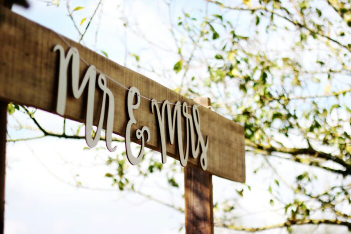 A rustic wooden wedding sign with white cursive lettering that reads "Mr & Mrs" hanging outdoors with green trees in the background.